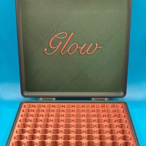 May include: A dark green storage case with copper-colored hinges and a copper-colored interior grid featuring many circular compartments. The inside of the lid displays the word Glow in a copper-colored cursive script font.
