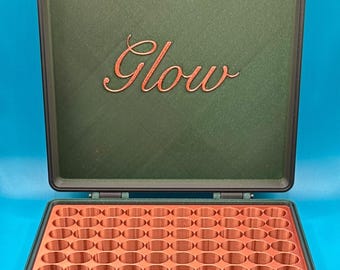 May include: A dark green storage case with copper-colored hinges and a copper-colored interior grid featuring many circular compartments. The inside of the lid displays the word Glow in a copper-colored cursive script font.