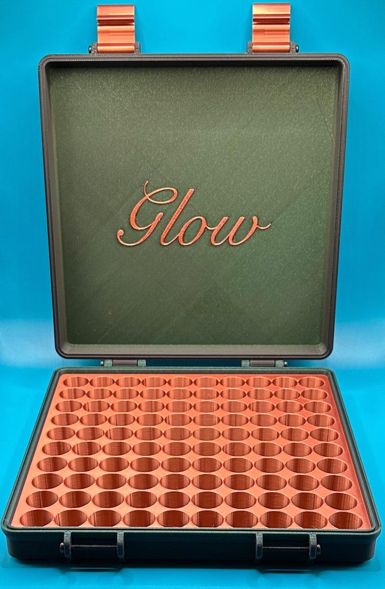 May include: A dark green storage case with copper-coloured hinges and a copper-coloured interior grid featuring many circular compartments. The inside of the lid displays the word Glow in a copper-coloured cursive script font.