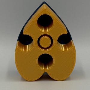 May include: A gold and navy blue, heart-shaped object with multiple circular openings. The object has a textured surface and a central circular feature. The design is symmetrical, with a rounded bottom and a pointed top.