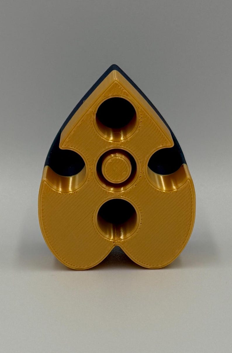 May include: A gold and navy blue, heart-shaped object with multiple circular openings. The object has a textured surface and a central circular feature. The design is symmetrical, with a rounded bottom and a pointed top.