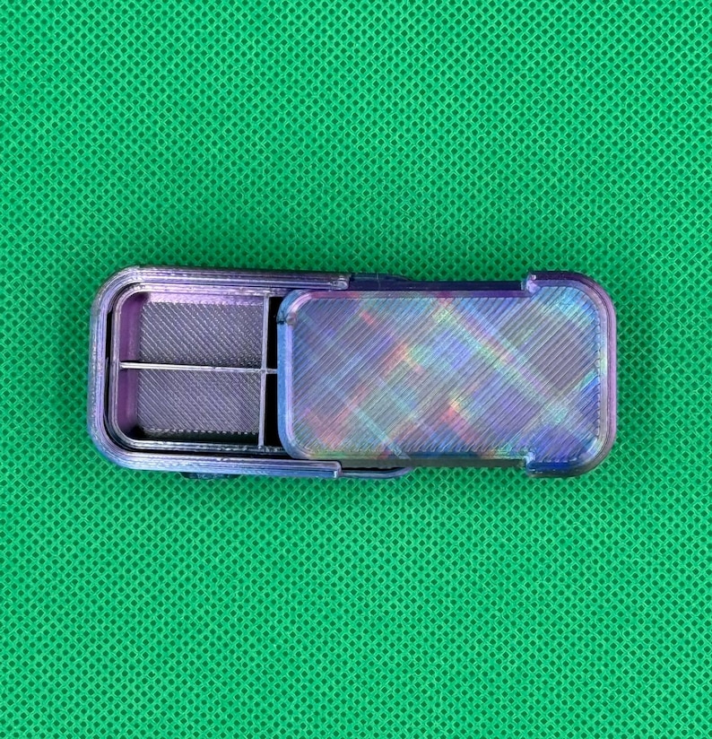 May include: A small, rectangular, iridescent pill case with a hinged lid. The case is divided into four compartments and has a textured, multi-colored finish. The open lid reveals the interior compartments, designed for organizing medication or small items.