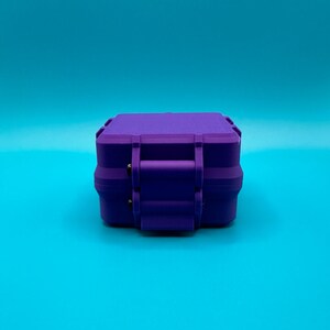 May include: A small, rectangular, purple hard-shell storage case with a secure latch mechanism on the front. The container features a matte finish and a structured, boxy design with reinforced edges, presented from a direct front-facing angle.