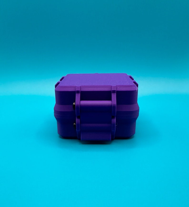 May include: A small, rectangular, purple hard-shell storage case with a secure latch mechanism on the front. The container features a matte finish and a structured, boxy design with reinforced edges, presented from a direct front-facing angle.