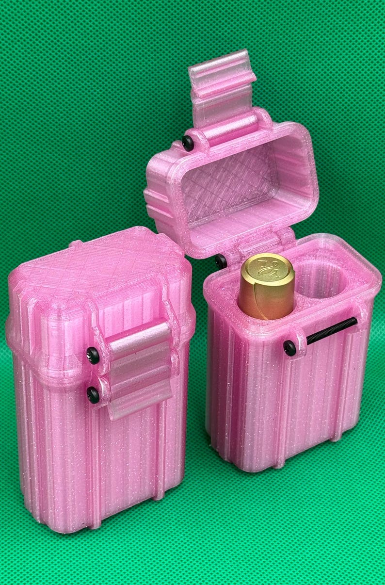 Puede incluir: Dos estuches de almacenamiento rectangulares de color rosa brillante con crestas verticales y pasadores de bisagra negros. Un estuche est&aacute; cerrado, mientras que el otro est&aacute; abierto, revelando un objeto cil&iacute;ndrico dorado.