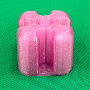 May include: A translucent pink, glitter-infused 3D printed object with a square-like shape featuring four rounded corners and deep, vertical channels carved into each side, creating a cross-like profile when viewed from above.