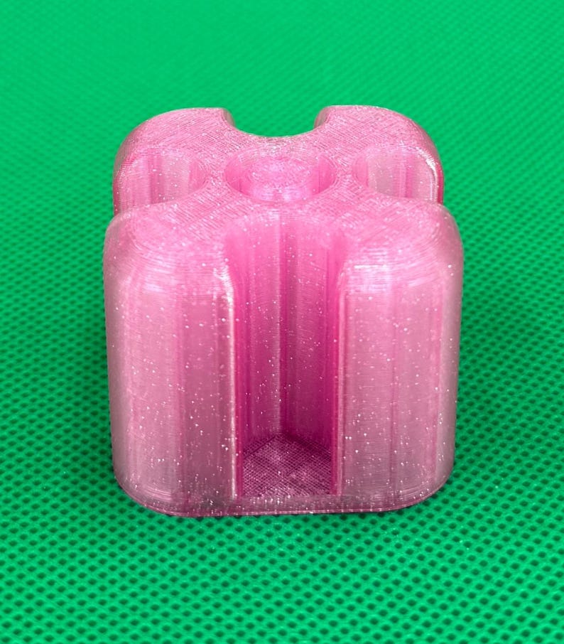May include: A translucent pink, glitter-infused 3D printed object with a square-like shape featuring four rounded corners and deep, vertical channels carved into each side, creating a cross-like profile when viewed from above.