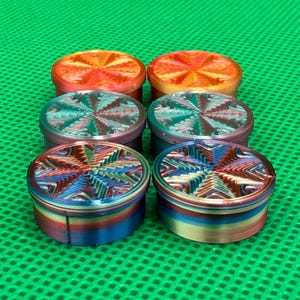 May include: Six cylindrical containers arranged in two rows of three. Each container features a vibrant, multi-colored gradient finish with a textured, star-like geometric pattern on the lid. The colors shift between orange, red, blue, and purple hues.
