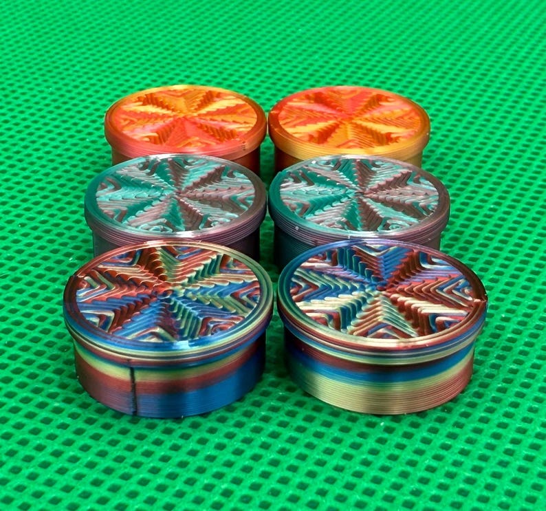 May include: Six cylindrical containers arranged in two rows of three. Each container features a vibrant, multi-colored gradient finish with a textured, star-like geometric pattern on the lid. The colors shift between orange, red, blue, and purple hues.