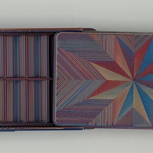May include: Open rectangular box with a hinged lid. The interior has compartments and a striped pattern. The lid features a starburst design in red, blue, and tan hues. The box is made of a material with a textured appearance.