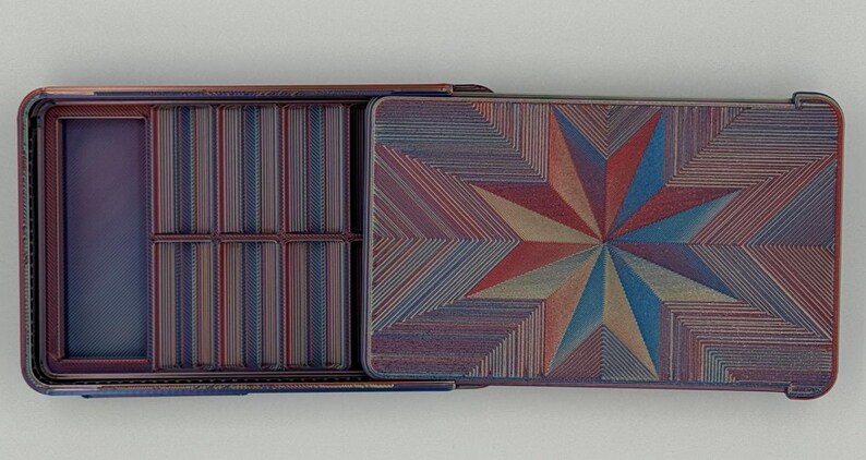 May include: Open rectangular box with a hinged lid. The interior has compartments and a striped pattern. The lid features a starburst design in red, blue, and tan hues. The box is made of a material with a textured appearance.