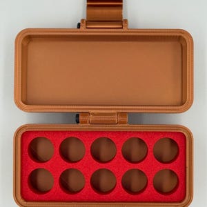 May include: A rectangular copper-colored plastic storage case with a hinged lid. The interior base features a bright red foam insert with ten circular cutouts arranged in two rows of five, designed for holding small cylindrical items.
