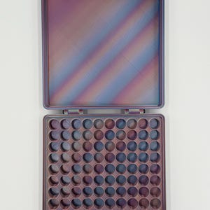 May include: A square, open storage case featuring a gradient of iridescent purple, blue, and copper tones. The interior base contains a grid of eighty-one circular compartments, while the lid is smooth with a matching multi-colored finish.