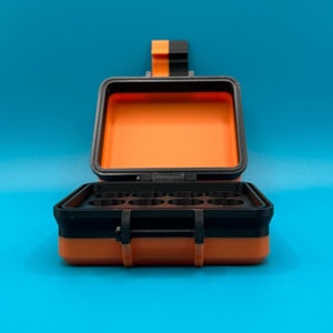 May include: A rectangular storage case with a black and orange exterior and a hinged lid. The interior features a black insert with eight circular compartments. The case includes a black and orange latch mechanism on the front and top.