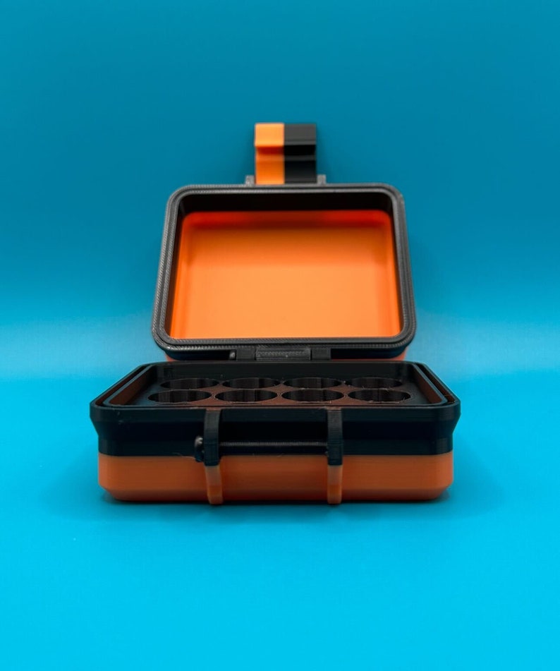 May include: A rectangular storage case with a black and orange exterior and a hinged lid. The interior features a black insert with eight circular compartments. The case includes a black and orange latch mechanism on the front and top.
