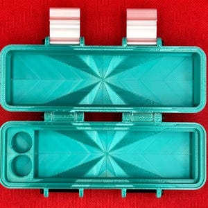 以下が含まれることがあります： ヒンジ付きの蓋と2つの銀色の金属製ラッチを備えた、青緑色の長方形の収納ケース。内部の両方のセクションの底にはスターバースト模様があり、下部トレイの左側には2つの円形のコンパートメントがあります。