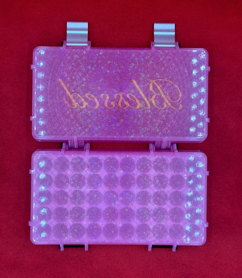 May include: A rectangular pink glitter storage case with a hinged lid. The lid features the word Blessed in gold cursive script. The interior base contains a grid of circular compartments for small items. The case has two silver-coloured latches.
