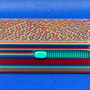May include: A rectangular storage box featuring a vibrant rainbow gradient finish with horizontal stripes on the sides and a complex geometric pattern on the lid. A small, bright teal sliding latch is centered on the front panel.