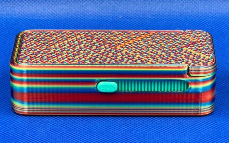 May include: A rectangular storage box featuring a vibrant rainbow gradient finish with horizontal stripes on the sides and a complex geometric pattern on the lid. A small, bright teal sliding latch is centered on the front panel.