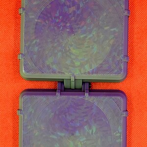 May include: A rectangular purple plastic case with a hinged design, featuring two identical sections. Each section has a circular holographic inlay with a concentric line pattern that reflects light in various iridescent colours.