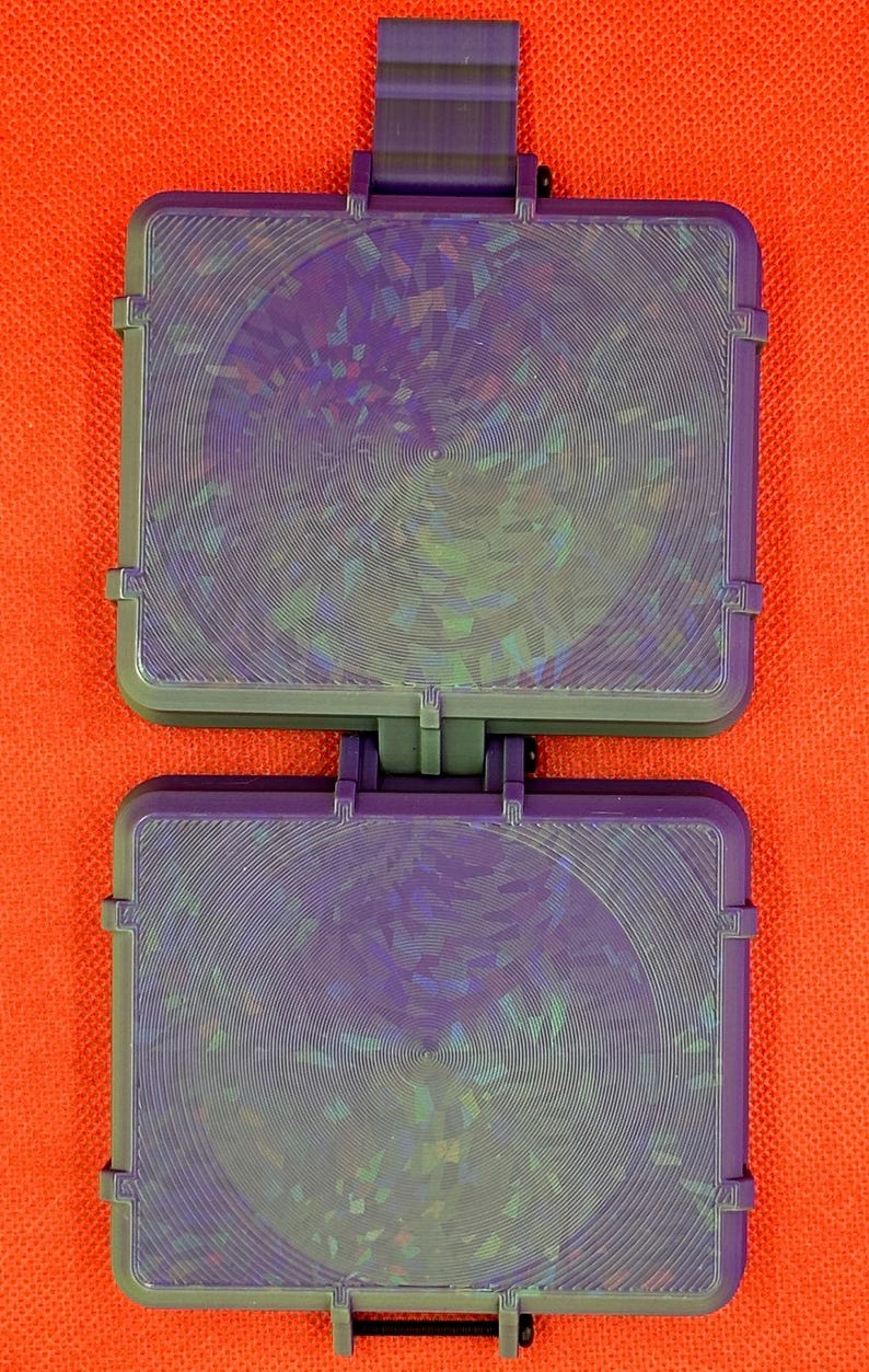 May include: A rectangular purple plastic case with a hinged design, featuring two identical sections. Each section has a circular holographic inlay with a concentric line pattern that reflects light in various iridescent colours.