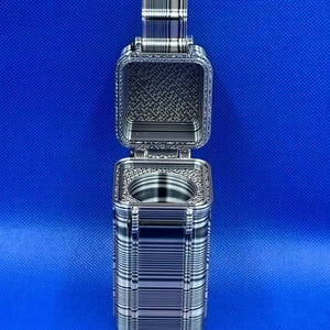 May include: A rectangular, metallic-toned container with a hinged lid that is open, revealing a circular interior cavity. The exterior features a distinct, repeating horizontal and vertical line pattern in shades of silver, gray, and black.
