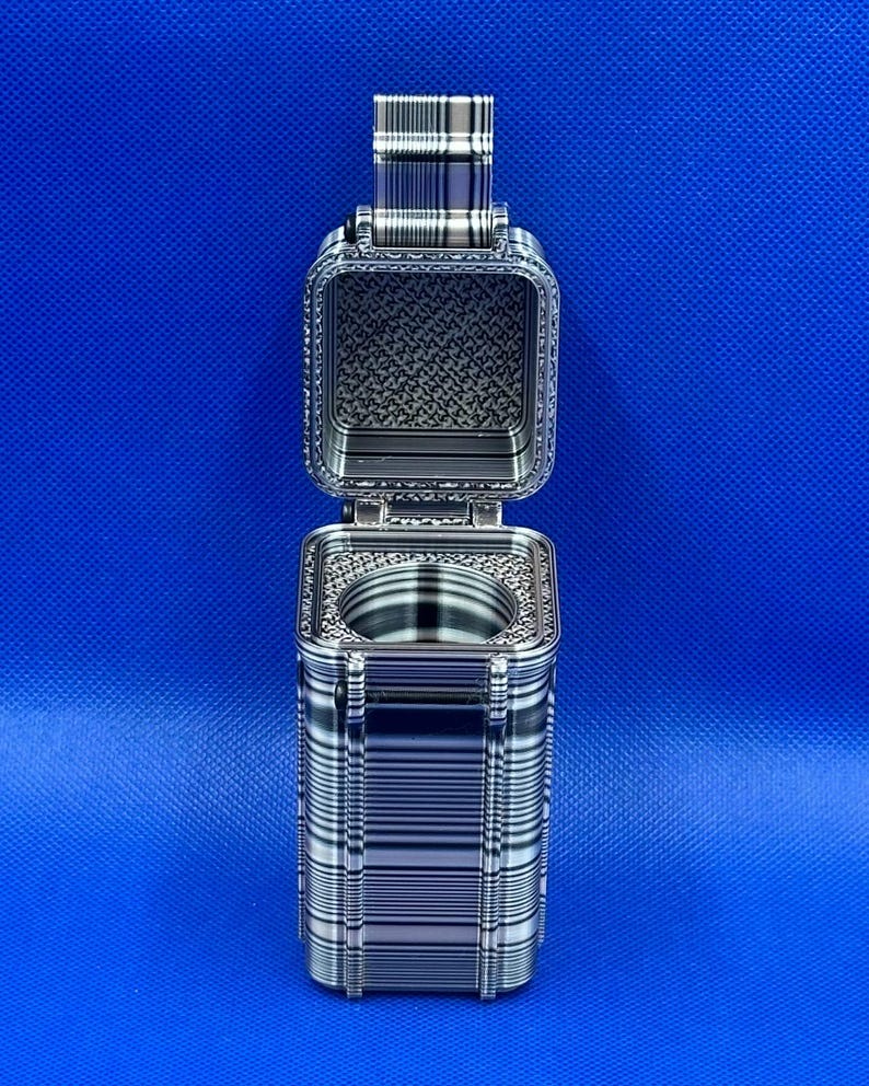 May include: A rectangular, metallic-toned container with a hinged lid that is open, revealing a circular interior cavity. The exterior features a distinct, repeating horizontal and vertical line pattern in shades of silver, gray, and black.