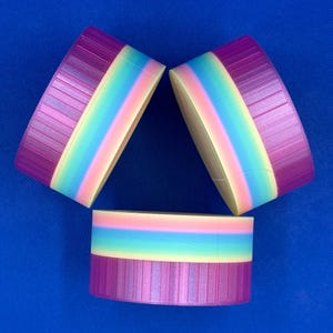 May include: Three cylindrical containers with ribbed purple lids and a rainbow-colored body featuring horizontal bands of pink, light blue, and yellow. The containers are arranged in a triangular formation against a solid blue background.