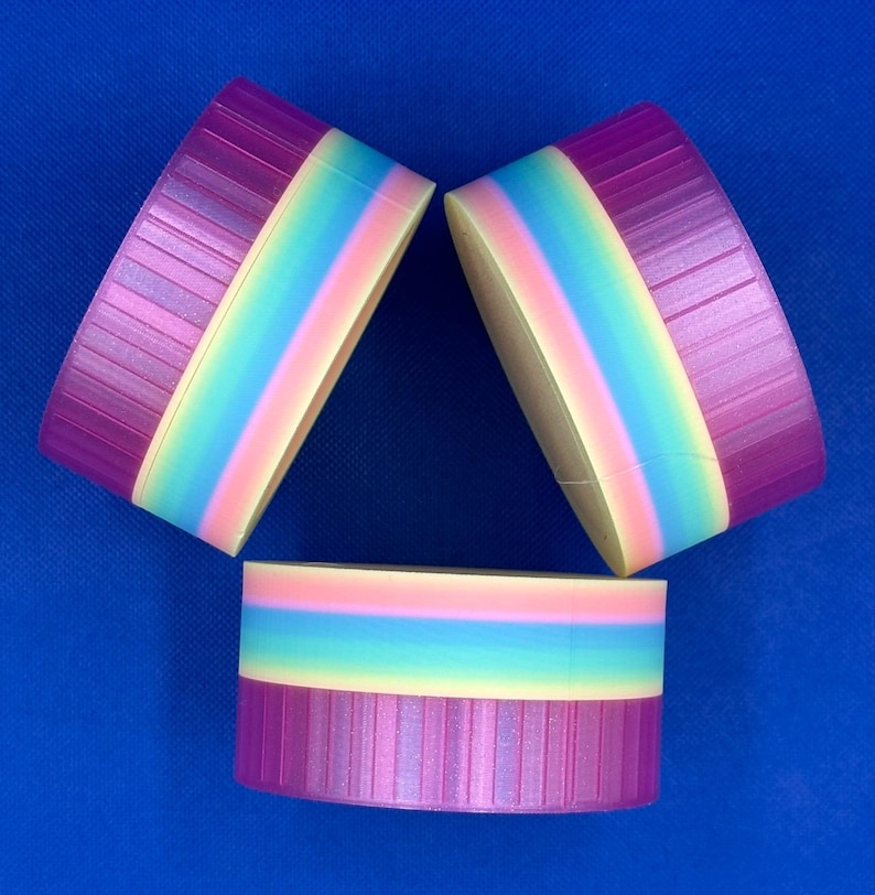 May include: Three cylindrical containers with ribbed purple lids and a rainbow-colored body featuring horizontal bands of pink, light blue, and yellow. The containers are arranged in a triangular formation against a solid blue background.