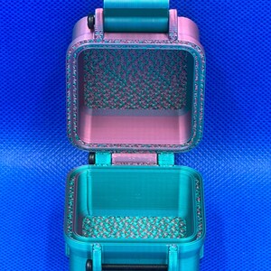 May include: A small, rectangular, open storage container with a hinged lid, featuring a dual-tone teal and pink color scheme. The interior surfaces display a textured, grid-like pattern, while the exterior has a smooth, layered finish.