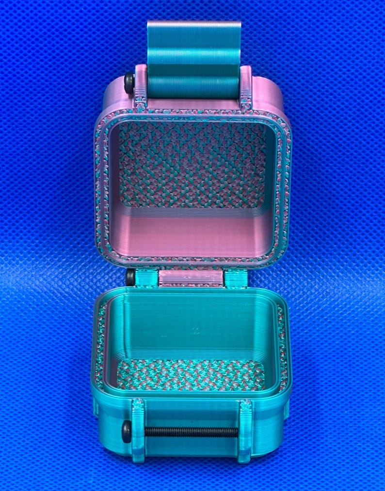May include: A small, rectangular, open storage container with a hinged lid, featuring a dual-tone teal and pink color scheme. The interior surfaces display a textured, grid-like pattern, while the exterior has a smooth, layered finish.
