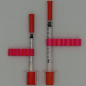 May include: Two syringes with orange caps and plungers, held by bright pink holders. The syringes are clear with black measurement markings. The holders have a series of small, evenly spaced ridges. The background is a plain, light gray.