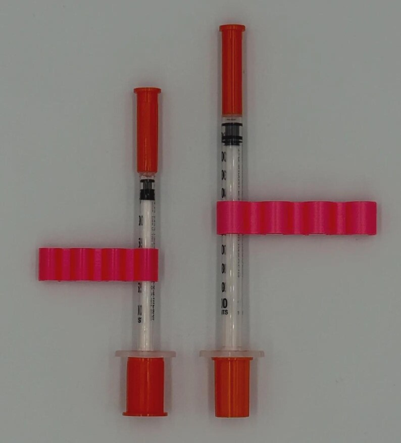 May include: Two syringes with orange caps and plungers, held by bright pink holders. The syringes are clear with black measurement markings. The holders have a series of small, evenly spaced ridges. The background is a plain, light gray.
