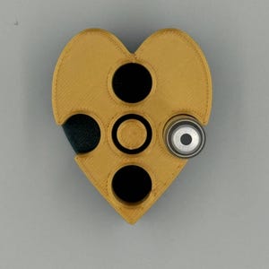May include: A heart-shaped, mustard-colored object with three black circular openings and a silver-colored cylindrical component. The object has a textured surface and appears to be 3D-printed.