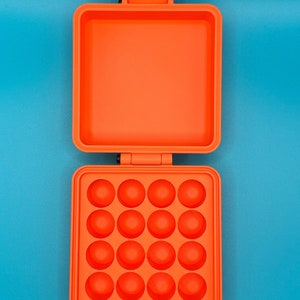 May include: A bright orange square plastic case with a hinged lid. The interior of the bottom section features a grid of sixteen circular indentations arranged in four rows of four, while the top lid interior is a smooth, flat square surface.