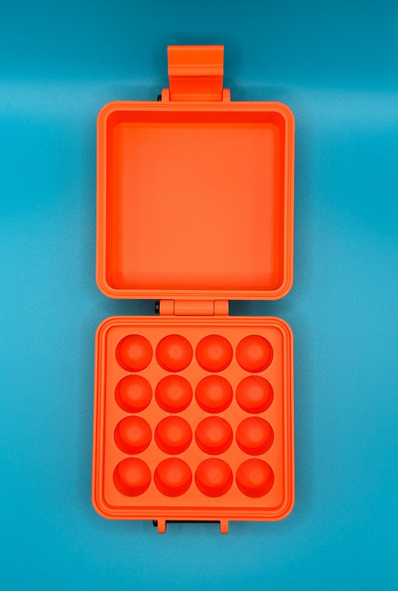 May include: A bright orange square plastic case with a hinged lid. The interior of the bottom section features a grid of sixteen circular indentations arranged in four rows of four, while the top lid interior is a smooth, flat square surface.