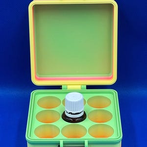 May include: A square, hinged storage box with a gradient rainbow exterior and a light green interior. The box features a grid insert with nine circular cutouts, one of which contains a small brown glass bottle with a white screw-top cap.