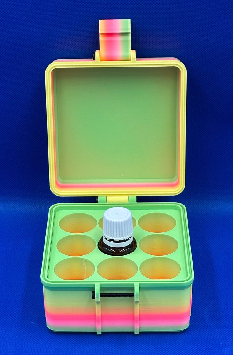 May include: A square, hinged storage box with a gradient rainbow exterior and a light green interior. The box features a grid insert with nine circular cutouts, one of which contains a small brown glass bottle with a white screw-top cap.