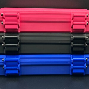 May include: A stack of three rectangular storage cases, each featuring two hinged locking latches on the front. The top case is bright pink, the middle case is black, and the bottom case is a vibrant blue, all with a textured finish.