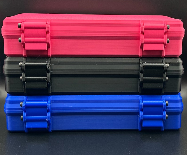 May include: A stack of three rectangular storage cases, each featuring two hinged locking latches on the front. The top case is bright pink, the middle case is black, and the bottom case is a vibrant blue, all with a textured finish.
