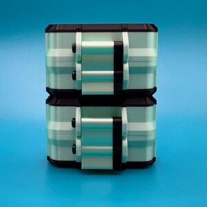 Puede incluir: Dos cajas de almacenamiento rectangulares apiladas con un cuerpo de color verde menta claro, bordes negros y mecanismos de cierre negros en la parte frontal. Cada caja tiene un dise&ntilde;o modular estructurado con costuras visibles.