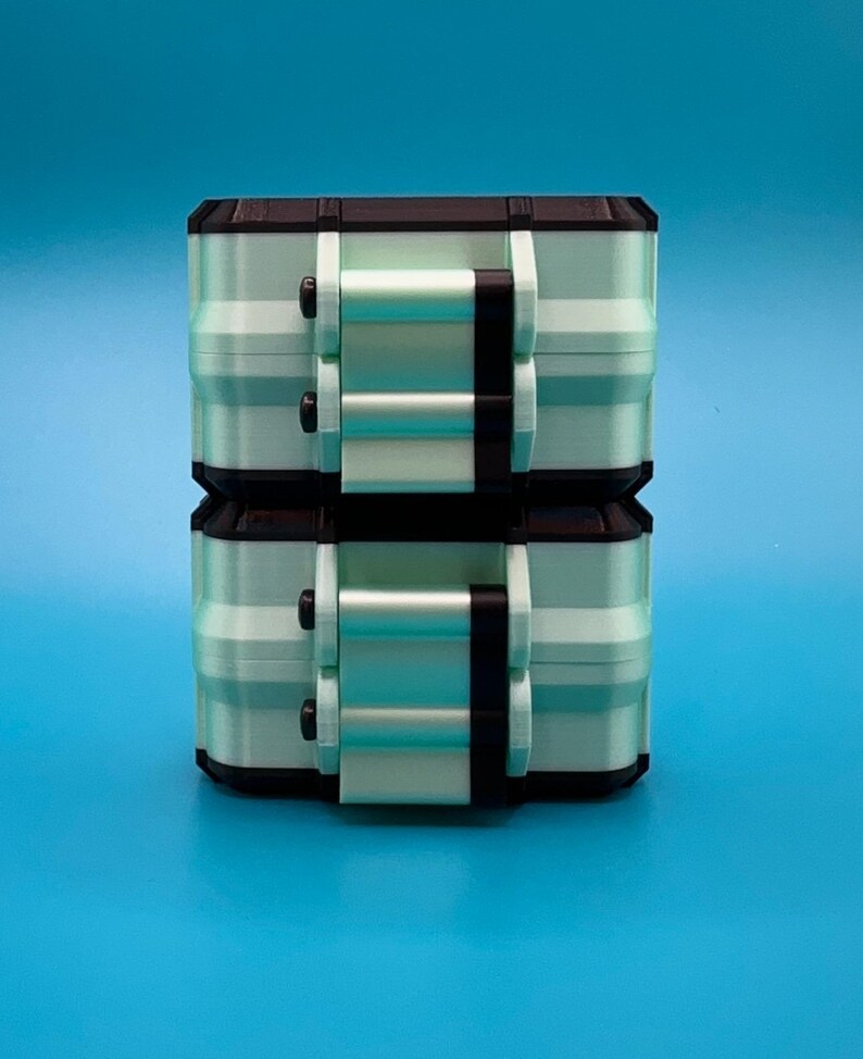 Puede incluir: Dos cajas de almacenamiento rectangulares apiladas con un cuerpo de color verde menta claro, bordes negros y mecanismos de cierre negros en la parte frontal. Cada caja tiene un dise&ntilde;o modular estructurado con costuras visibles.