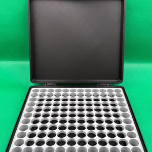 May include: Black plastic storage case with a white foam insert containing 100 round compartments. The case is open and the lid is propped up.