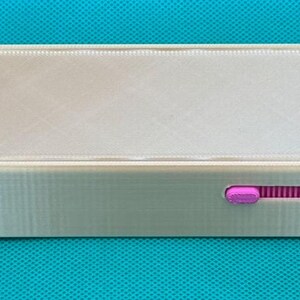 May include: A rectangular, light gray plastic storage box with a sliding lid. The interior features a bright pink compartment on the left side. A small pink sliding mechanism is visible on the side of the box, allowing the lid to open.