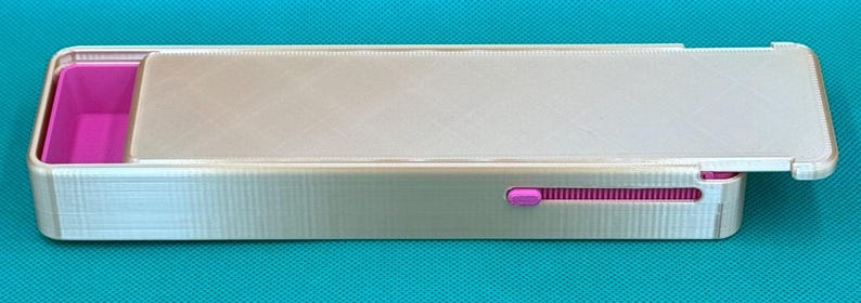 May include: A rectangular, light gray plastic storage box with a sliding lid. The interior features a bright pink compartment on the left side. A small pink sliding mechanism is visible on the side of the box, allowing the lid to open.