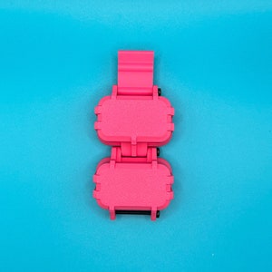 May include: A bright pink, rectangular, hinged storage case featuring two connected compartments with a vertical strap attachment. The exterior has a textured, matte finish with small black hardware accents visible at the hinges and joints.