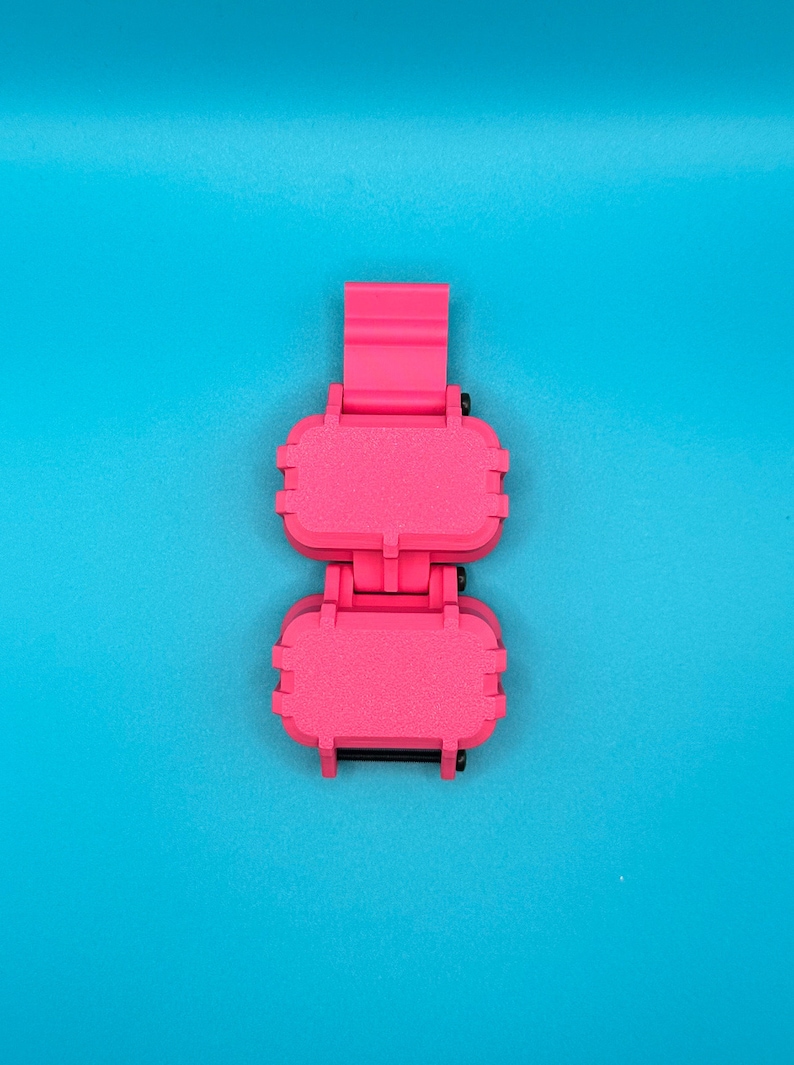 May include: A bright pink, rectangular, hinged storage case featuring two connected compartments with a vertical strap attachment. The exterior has a textured, matte finish with small black hardware accents visible at the hinges and joints.