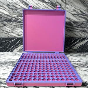 May include: An open, rectangular, two-tone case with a pink interior and a purple exterior. The case is divided into a grid of small, circular holes. The case is sitting on a marble surface.