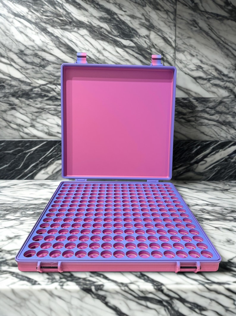 May include: An open, rectangular, two-tone case with a pink interior and a purple exterior. The case is divided into a grid of small, circular holes. The case is sitting on a marble surface.
