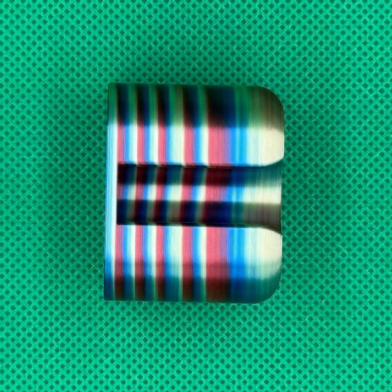 May include: A cylindrical metal object featuring a vibrant, multi-colored anodized finish with alternating vertical stripes of deep blue, magenta, and metallic silver. The piece has a smooth, polished surface and a rectangular cutout section.